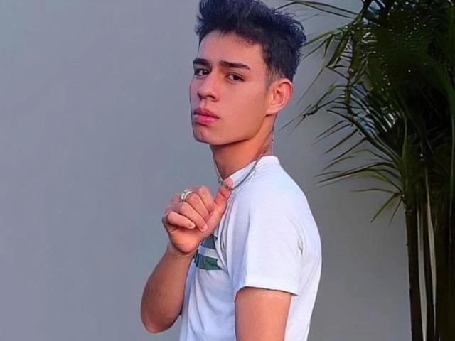 AngelHub, un joven con cabello corto y oscuro, tirando de un collar con una cadena alrededor de su cuello, vistiendo una camiseta blanca, con los ojos cerrados, la cabeza inclinada hacia atrás, con una expresión seria..