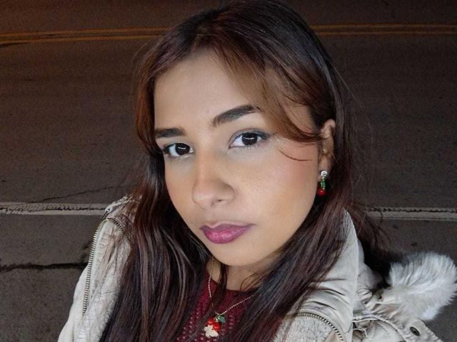 MariamLara hat braunes Haar, hellbraune Haut und trägt rosafarbenen Lippenstift mit grünen Ohrringen, ein rotes Hemd und eine beige Jacke. Ihr Hintern ist nicht sichtbar..