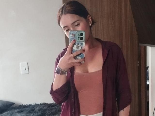 SarahShinny with red-brown hair, wearing a maroon shirt over a pink tank top, taking a mirror selfie with a phone, showing her small tits and subtle ass.
