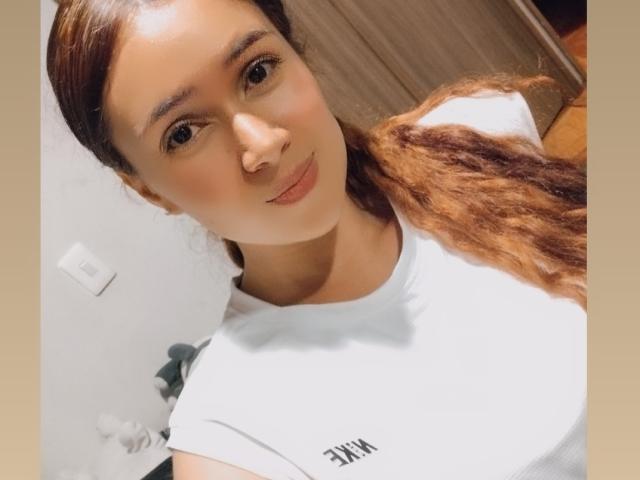 SarahShinny met bruin, golvend haar in een staart, draagt een witte blouse, glimlacht naar de camera, haar gezicht en borst zijn zichtbaar, zacht licht, lichtrode huid..
