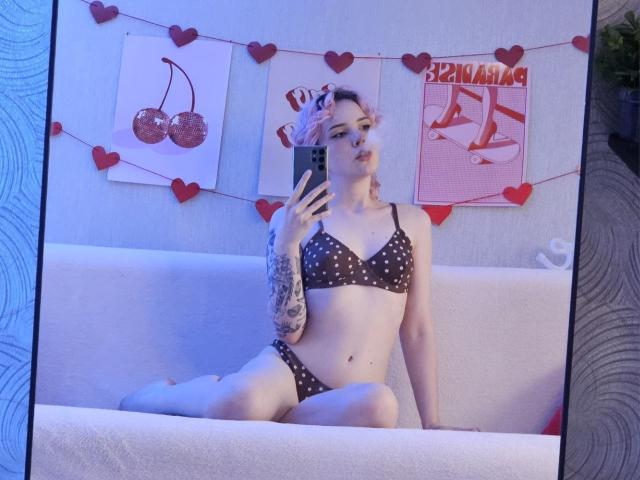ShellyLove69 met roze haar, een bikini met stippen, een getatoeëerde arm en kleine borsten, zittend op een witte bank, een spiegelzelfportret makend, met blauw licht, rode hartdecoraties en kersenkunst..