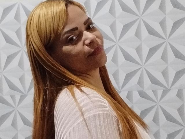 DeborahSweetX, avec de longs cheveux blonds, une peau légèrement brune et un sourire discret, porte un haut rose transparent et regarde par-dessus son épaule. Ses seins sont légèrement visibles à travers le haut..