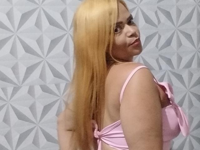 DeborahSweetX, avec de longs cheveux blonds, un soutien-gorge rose, et un regard de côté, montrant son dos et ses seins, debout devant un mur avec un motif géométrique..
