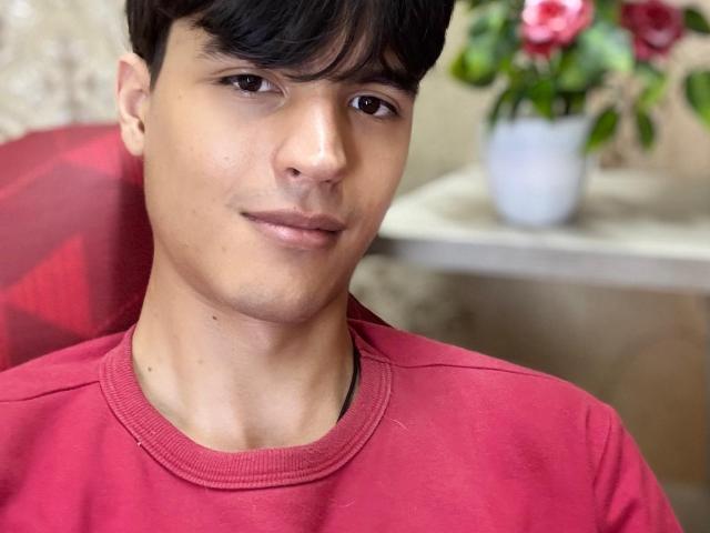 TylerMulber, medium skin tone, short black hair, brown eyes, slight smile, wearing red shirt, nose is small, pouty lips, small visible mole on right cheek, white flower vase in background.