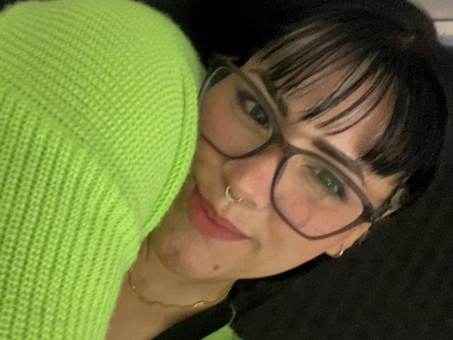 AlejaRossi with green sweater, black hair, glasses, nose ring, and gold necklace, smiling at camera, showing her tits slightly. Her green eyes are visible.