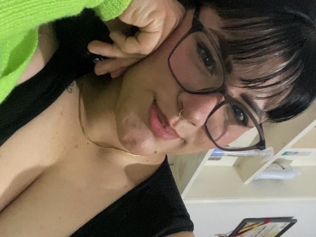 AlejaRossi con gafas, cabello negro, piercing en la nariz y una blusa verde, sonriendo, con la mano en la mejilla, mostrando sus senos, rostro ligeramente sucio, en un selfie..