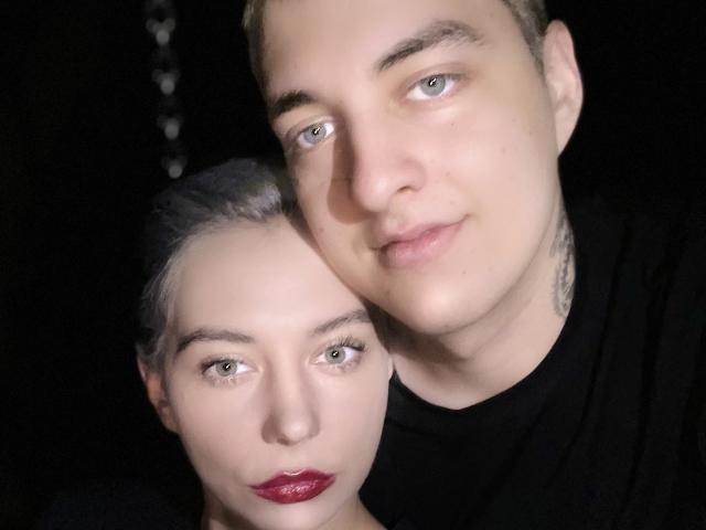 BlondIce, con cabello rubio, camisa negra y piel pálida, sosteniendo la cabeza de una mujer con cabello plateado, lápiz labial rojo y piel pálida, que está de pie detrás de ella..