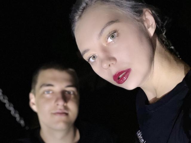 BlondIce en een man met kort bruin haar, beiden in zwarte shirts, kijken naar de camera. BlondIce heeft rode lippenstift en grijs haar, de man heeft blauwe ogen..