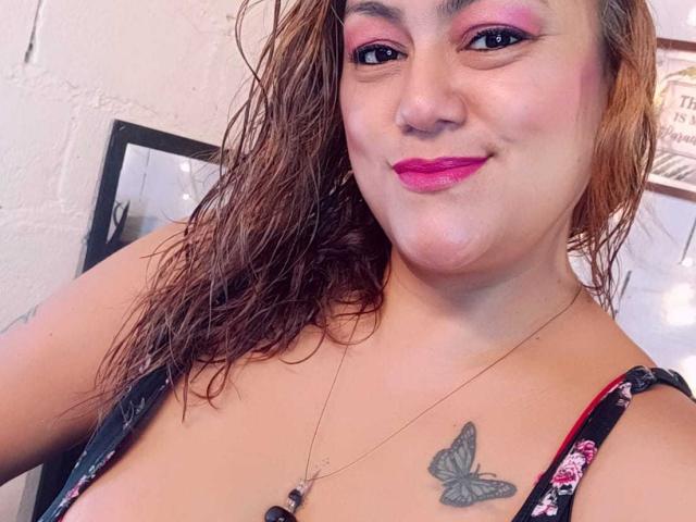 MissRosarioHot se tient debout, chaussée de talons blancs, et porte des sous-vêtements noirs à fines bretelles, exposant ses fesses nues et ses tatouages, avec une jambe levée, devant un mur en briques blanches..