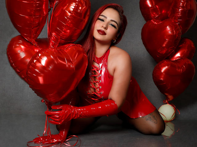 Sophia, een vrouw met rood haar, gekleed in een glanzende rode latex outfit, zittend op een gouden stoel, benen gekruist, handen in haar haar, kerstboom aan de rechterkant..
