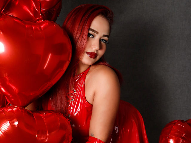 SophiaBlakes with red hair, wearing a shiny red latex outfit, bending over a gold chair, touching a decorated Christmas tree, showing her ass, tattoo on her left arm.