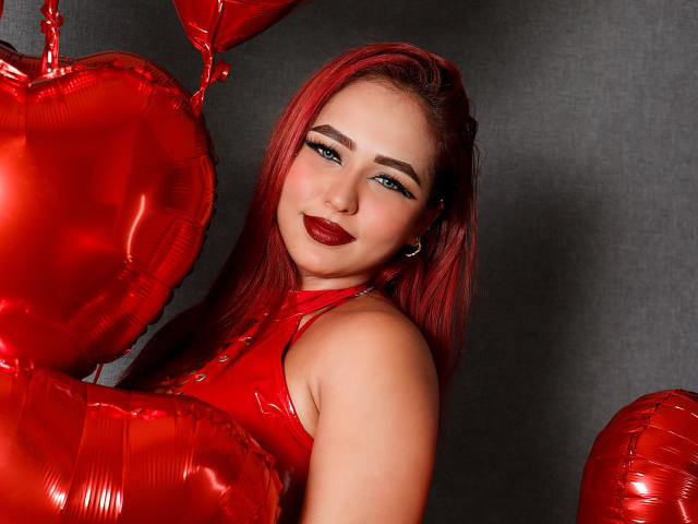 SophiaBlakes vestindo uma roupa de látex vermelha brilhante, luvas vermelhas e balões em formato de coração vermelhos, ajoelhada, segurando um balão, batom vermelho, tatuagem na coxa, fundo cinza, expressão sedutora e confiante..
