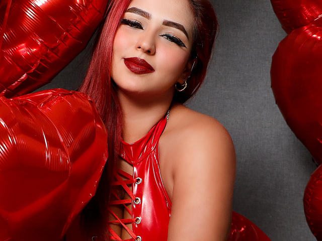 SophiaBlakes with red hair, wearing shiny red latex, holding red heart balloons, has red lipstick, and a tattoo on her right arm, looking seductively at the camera.