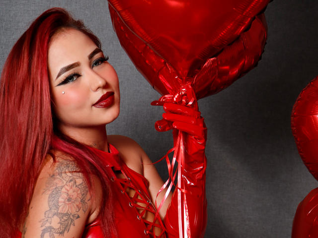 Sophia, con cabello rojo, vestida con una brillante parte superior de látex rojo, sosteniendo globos rojos con forma de corazón, tiene ojos azules, lápiz labial rojo oscuro y una sonrisa juguetona, mirando a la cámara..