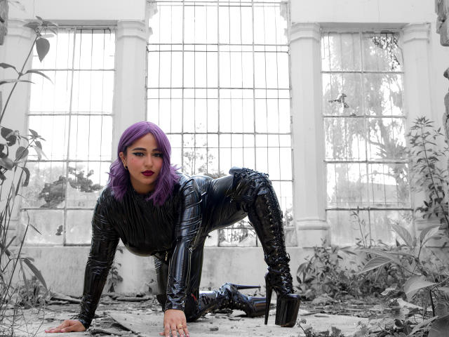 Sophia, com cabelo roxo, vestindo um macacão preto, justo, brilhante e de látex, encostada em uma parede branca, exibindo suas nádegas e olhando por cima do ombro com uma expressão confiante..