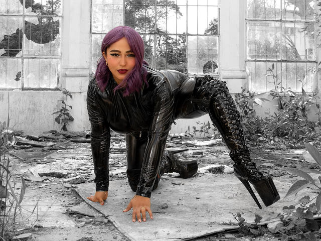 SophiaBlakes with purple hair, wearing a black latex bodysuit and boots, squatting between white pillars. She has large tits, a cute face, and dark lipstick.