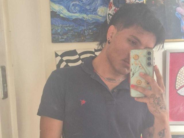 BillyReynols with short black hair, wearing a dark polo shirt, holding a phone with a floral case, showing tattoos on his hand and ear, taking a mirror selfie.