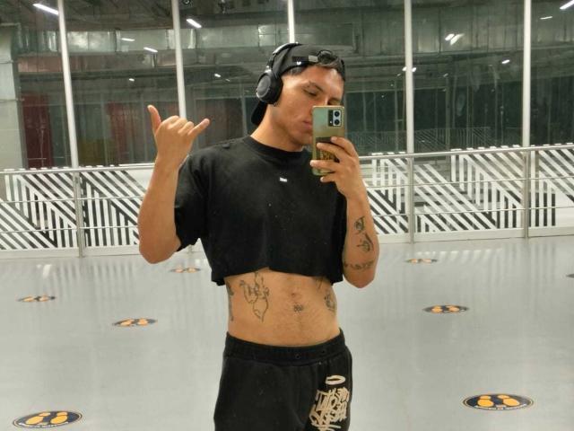 BillyReynols with tattoos, wearing a black crop top and headphones, showing a middle finger, holding a phone, standing in a gym with a gray floor.