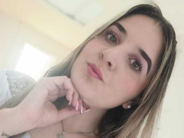 KatyWowBlue a la peau claire, les yeux marron et du rouge à lèvres rose. Elle se tient le menton avec ses ongles roses parfaitement manucurés, et regarde l'appareil photo avec une légère moue..