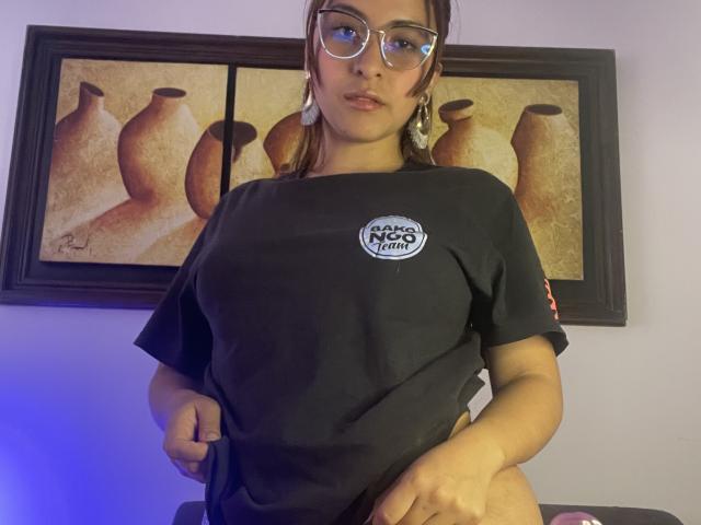 AlannaN with glasses, wearing a black t-shirt, slightly lifting it to reveal her black panties, showing her butt. Brown hair, earrings, and a serious expression.
