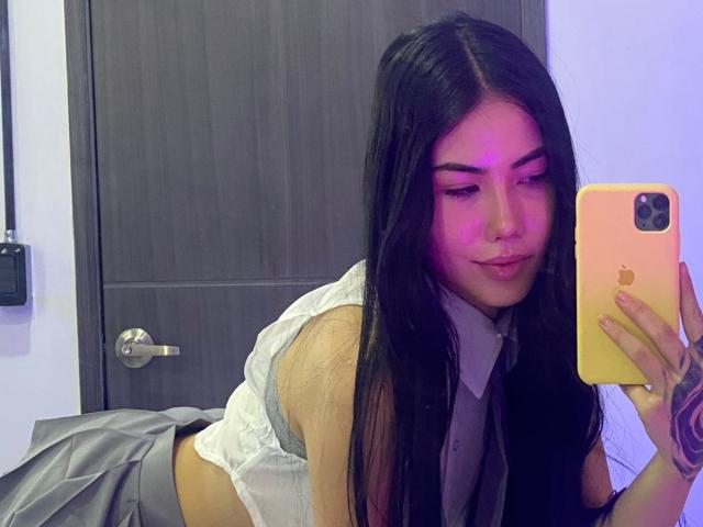 VictoriaLys, com longos cabelos pretos, vestindo uma camisa branca e uma saia cinza, deitada, tirando uma selfie no espelho com um iPhone com um tom rosado, mostrando seu rosto fofo e seios pequenos..