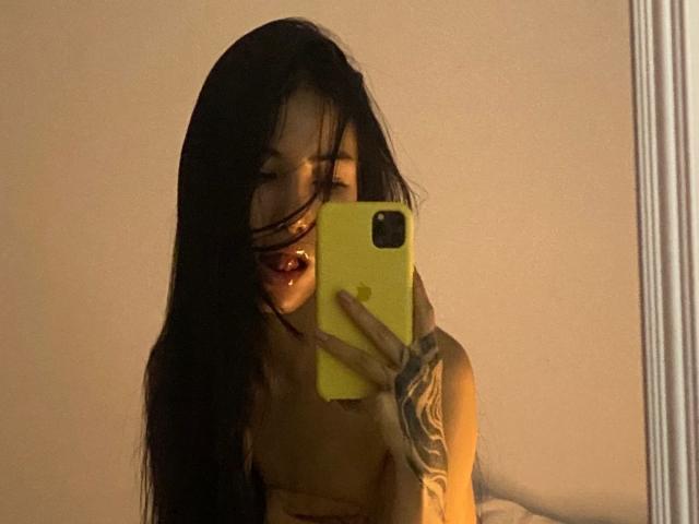VictoriaLys, avec de longs cheveux noirs, tenant un iPhone jaune, cachant ses petites seins avec sa main, tatouage sur son bras droit, selfie devant un miroir, la bouche légèrement ouverte..