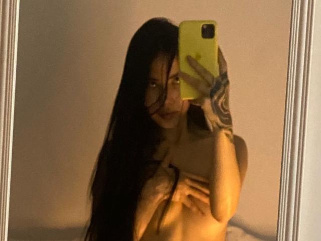 VictoriaLys mit langen, schwarzen Haaren, die ihre Brüste mit den Händen bedeckt, macht ein Selfie mit einem gelben Handy und zeigt dabei ein Tattoo auf ihrem rechten Arm..