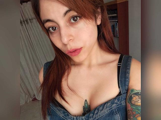 AdelaLove69, avec ses yeux marron, ses lèvres rouges et ses longs cheveux bruns, porte une robe en jean qui dévoile sa poitrine et a un tatouage vert sur le côté droit. Ses seins sont partiellement visibles..