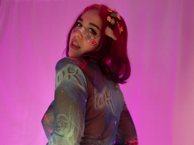 Toukauwu, com cabelo ruivo, vestindo uma blusa azul transparente, tem glitter no rosto, uma coroa de flores e orelhas de vaca, olhando para trás com uma expressão brincalhona. Luz rosa e roxa..
