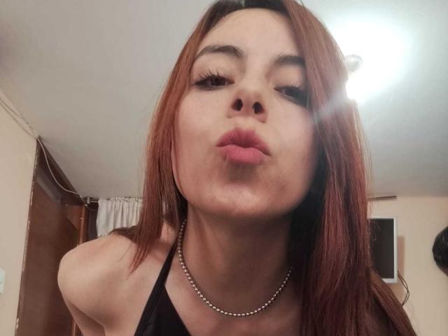 AdelaLove69, con le labbra tumefatte, indossa una maglietta nera e una collana d'argento, ha lunghi capelli rossi e fa un'espressione civettuola, guardando verso il basso la fotocamera..