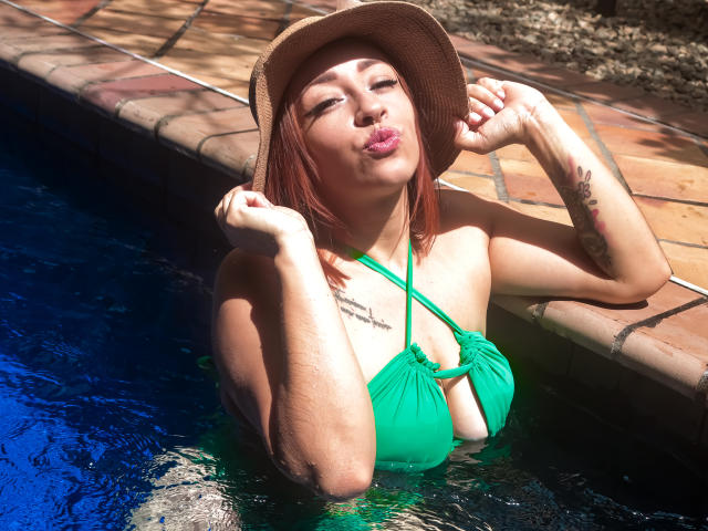 CharlotRey en maillot de bain vert, les seins au-dessus de l'eau, portant un chapeau, lèvres boudeuses, tatouage sur le bras droit, dans une piscine, lumière du soleil, eau bleue, bordure en briques..