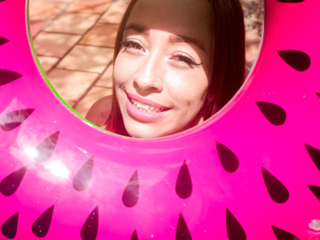 Het lachende gezicht van CharlotRey, met roze lippenstift en een neusring, is te zien door een roze, op watermeloen gemotiveerde opblaasbare vorm. Zonlicht accentueert haar bruine haar en witte eyeliner..