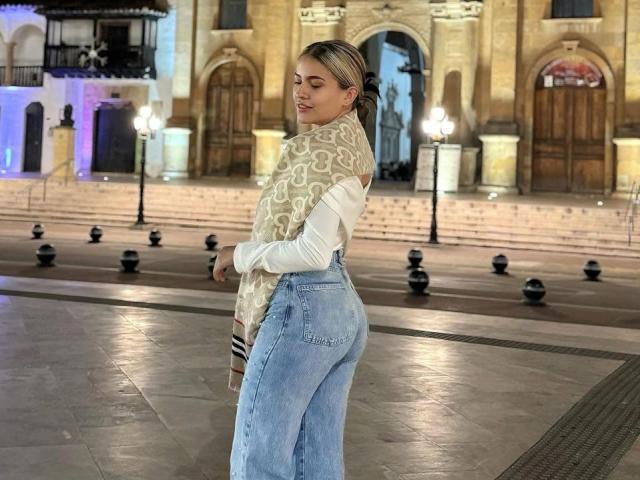 JasminBela, com cabelo loiro preso em um rabo de cavalo, usando um lenço bege estampado, camisa branca e jeans azuis de cintura alta, mostrando o bumbum, está em uma praça à noite..