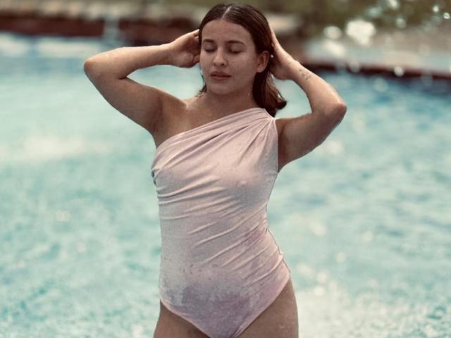 JasminBela usando um biquíni rosa claro de um ombro só, em pé em uma piscina, com os braços levantados e os olhos fechados, com gotas de água na pele, mostrando seus pequenos seios e nádegas molhadas..