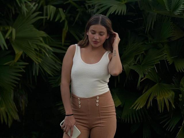 JasminBela met bruin haar, een wit tanktopje en beige, hoge broek, die een witte telefoon vasthoudt, naar beneden kijkt en voor donkergroene palmbladeren staat..