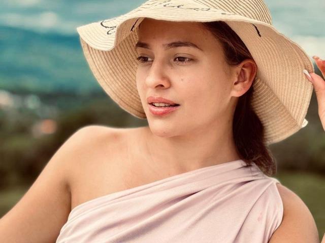 JasminBela, con la pelle color nocciola, gli occhi marroni e il naso piccolo, indossa un cappello beige e una maglietta rosa con una spalla scoperta, tenendo il bordo del cappello, con la bocca leggermente aperta..