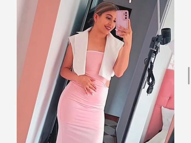 JasminBela, con i capelli biondi, un vestito rosa, un gilet bianco senza maniche, che tiene in mano il telefono, sta scattando un selfie allo specchio, mostrando i suoi fianchi formosi, è in piedi, sorride, in una stanza ben illuminata, la fotocamera è a destra..
