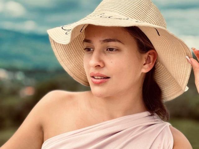 JasminBela, con piel morena, ojos marrones y labios rosados, lleva un sombrero de paja beige y una blusa rosa con un solo hombro, sosteniendo su sombrero. Los senos están parcialmente cubiertos por la blusa..