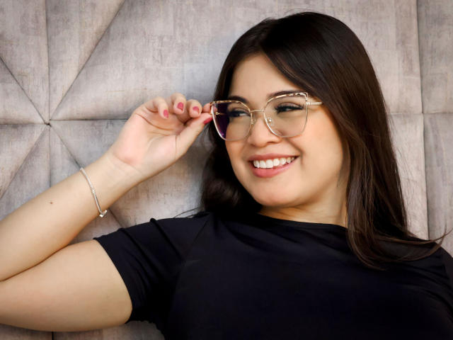 CamilaSofiia with glasses, black shirt, smiling, brown hair, holding glasses with pink nails, silver bracelet, cute, attractive, medium tits, light skin, relaxed, happy, casual.