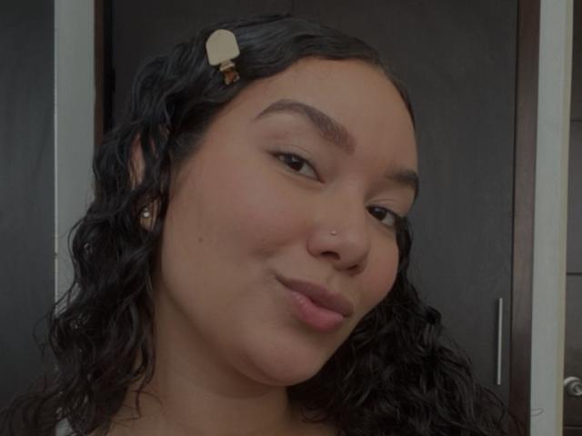 Ashlley69 hat lockiges, schwarzes Haar mit einer beigen Haarnadel, einen Nasenring und ein leichtes Lächeln, blickt zur Seite mit vollen Lippen und einem süßen, entspannten Gesicht..