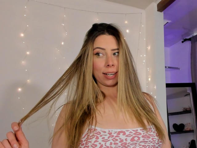 JennyLawry with long blonde hair, holding a strand, wearing a pink and white leopard print top, smiling, fair skin, and light makeup, in a softly lit room.