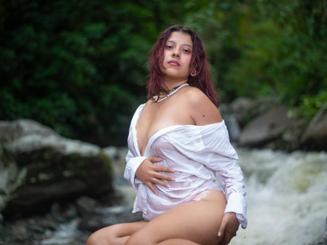 Coraline Hale, avec des cheveux roux, portant une chemise blanche ouverte qui expose ses seins, assise près d'un ruisseau, une main sur son ventre, un collier, et des sous-vêtements roses, regardant l'appareil photo..