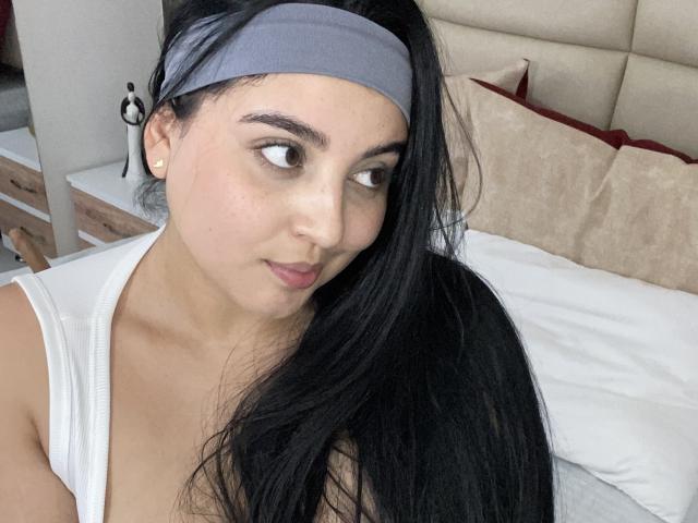 ShinyMurpy avec de grandes seins, portant un crop top blanc, un bandeau gris, des cheveux noirs, souriante, assise sur un lit, avec des oreillers blancs et beige, un sextoy sur le lit, un smartphone..