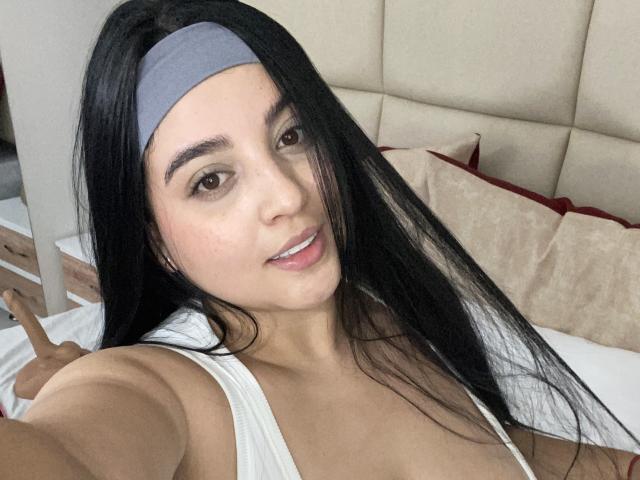 LilithHellsing with a colorful hat, white tee, and black thong, sitting on a bed, showing her round ass, wearing a thong, and smiling at the camera.