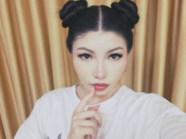 ViolettaLeroy with black double buns, wearing a white shirt, has her finger to her pink lips, looking seductively at the camera, with pale skin and dark eyes.