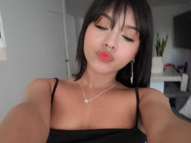 HannaCrow with pouty red lips, black spaghetti straps, small tits, brown hair, silver necklace, earrings, and nose ring, taking a selfie, eyes closed, in a soft-lit room.