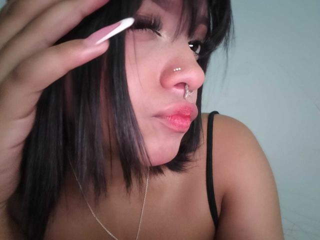 HannaCrow, com cabelo preto, piercings no nariz e batom rosa, piscando, mão com unhas brancas longas, usando uma alça preta, com o peito à mostra, pele clara, close-up..