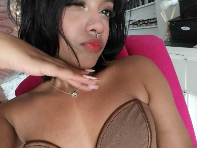 HannaCrow, con i capelli neri, un piercing al naso e rossetto rosso, che fa l'occhiolino, toccandosi il collo, indossa un top marrone senza spalline, mostrando il seno, seduta su una sedia rosa..