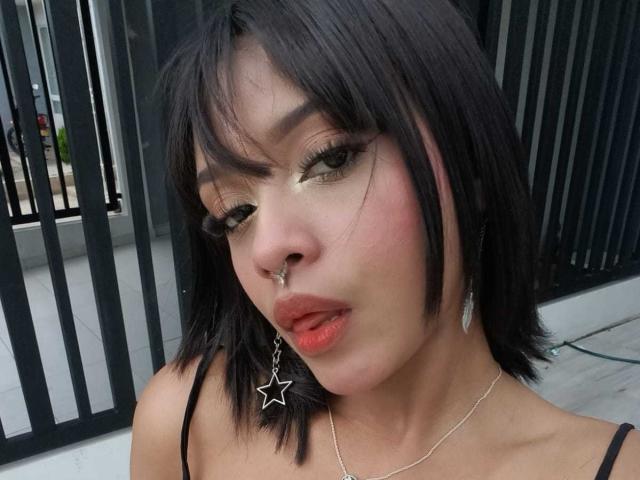 HannaCrow tem cabelo preto na altura dos ombros, um piercing no nariz, brincos em formato de estrela e delineador grosso. Seus lábios rosados estão ligeiramente entreabertos. Ela está vestindo uma blusa preta com uma alça fina..