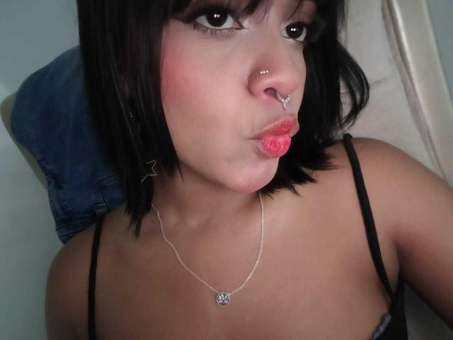 HannaCrow, con capelli neri, piercing al naso e al labbro, che fa la smorfia con le labbra rosse, indossa una canottiera nera, una collana d'argento e, sullo sfondo, una giacca di jeans..